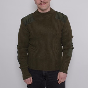 Może przedstawiać: Oliwkowozielony sweter z prążkowanej dzianiny z wzmocnionymi naszywkami na ramionach i łokciach. Sweter ma wysoki, okrągły dekolt i długie rękawy. Osoba ma na sobie czarne dżinsy.