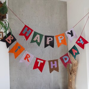Puede incluir: Una guirnalda de tela que deletrea HAPPY BIRTHDAY con banderines individuales de varios colores, incluyendo rojo, naranja, verde, negro y blanco. Cada letra está aplicada sobre los banderines usando diferentes telas estampadas y lisas.