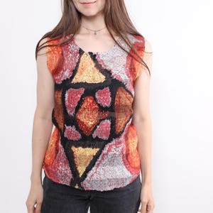 Puede incluir: Top sin mangas con un estampado geométrico en tonos rojos, naranjas, negros y dorados. El top tiene un escote redondo y está hecho de una tela texturizada. La modelo lleva vaqueros negros.