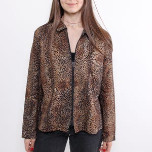 May include: A long-sleeved, collared shirt with a leopard print pattern in shades of brown and tan. The shirt has a full-length black zipper down the front. The person is wearing black jeans.