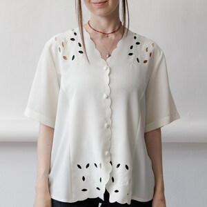 Puede incluir: Una blusa de manga corta en color blanco roto con borde frontal festoneado, cuello en V y botones a juego forrados en tela. La prenda presenta patrones de bordado floral recortados a lo largo de los hombros y el dobladillo inferior.