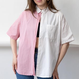 May include: A short-sleeved button-down shirt featuring a split-color design with a solid pink panel on the right side and a solid white panel on the left side. The shirt includes a standard collar, a front button placket, and a single chest pocket.