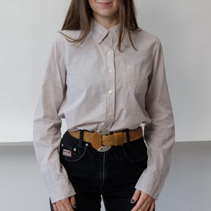 May include: A long-sleeved button-down shirt with a fine beige and white grid pattern, paired with black denim jeans and a tan suede belt featuring a large, rounded silver-toned metal buckle. The shirt includes a single chest pocket.