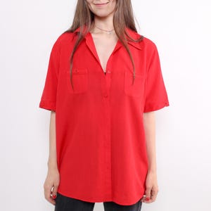Puede incluir: Una camisa roja brillante de manga corta con botones y dos bolsillos en el pecho. La camisa tiene un cuello clásico y está hecha de una tela ligera. Las mangas están dobladas. La camisa es de color liso y de estilo vintage.