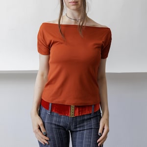 Peut inclure: Un haut couleur rouille à épaules dénudées avec des manches courtes et des détails froncés aux épaules, associé à une large ceinture en velours rouge avec une boucle dorée et un pantalon bleu foncé à fines rayures verticales.