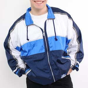May include: A color-blocked windbreaker jacket featuring horizontal panels of white, bright blue, and dark navy blue. The jacket has a full-length front zipper, a high collar with drawstring cords, and long sleeves with dark navy blue accents.