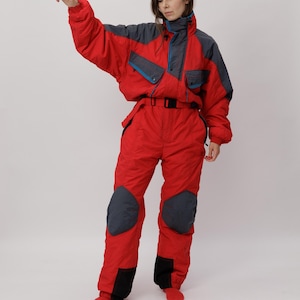 May include: A red and charcoal grey one-piece snowsuit featuring a high collar, long sleeves with elastic cuffs, a waist belt with a black buckle, and reinforced charcoal grey patches on the knees and lower legs with blue accent piping details.