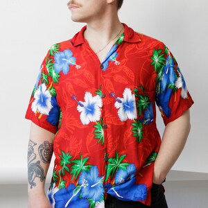 May include: Red short-sleeve button-down shirt with a floral pattern featuring blue and white hibiscus flowers and green leaves. The shirt has a classic collar and is worn with black pants. The shirt is a vibrant piece of clothing.