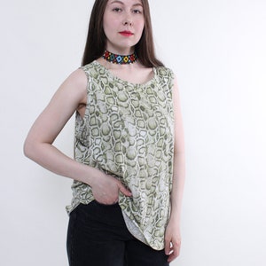 Könnte beinhalten: Eine Frau trägt ein ärmelloses Oberteil mit Schlangenhautmuster in Grün und Weiß mit einer bunten Perlenkette und einer schwarzen Jeans.