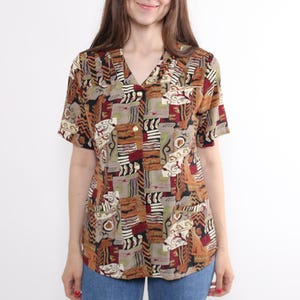 May include: Short-sleeved button-down shirt with a V-neck collar. The shirt features a geometric pattern in shades of brown, red, beige, and olive green. The shirt is paired with blue jeans. The model is smiling.