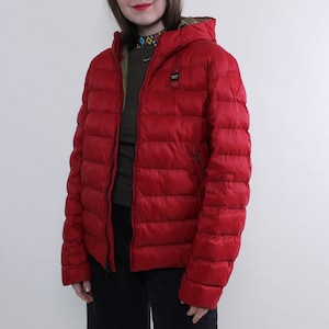 Könnte beinhalten: Rote Steppjacke mit Kapuze. Die Jacke hat ein gestepptes Design und einen Reißverschluss. Das Model trägt ein dunkelgrünes Hemd darunter. Der Hintergrund ist eine schlichte weiße Wand.