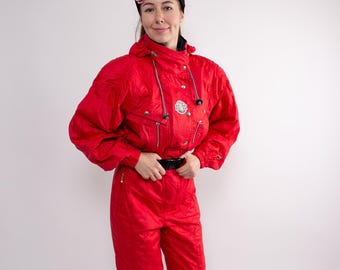 May include: A bright red ski suit with a matching hood and pants. The one-piece suit has a black belt, multiple zippers, and a small logo on the chest. The person is wearing black shoes and pink ski goggles.