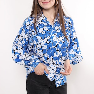 Può includere: Una camicetta blu con stampa floreale con dettagli bianchi e neri. La camicia a maniche lunghe ha un colletto e si abbottona sul davanti. La modella indossa jeans neri e orecchini a cerchio dorati.