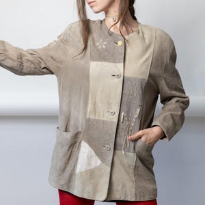 May include: A beige and taupe patchwork suede jacket featuring a round neckline, long sleeves, and a front button closure. The garment includes two side pockets and decorative stitching details on the front panels with a small gold-tone flower pin.