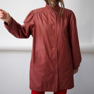 May include: A terracotta-coloured jacket featuring a high collar, long sleeves with gathered shoulders, and a front button closure. The garment includes two side pockets and a straight hemline, providing a structured look with a smooth finish.