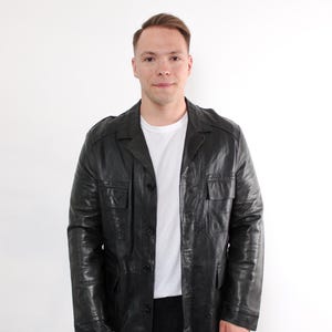 Op de afbeelding: Zwarte leren jas met een knoopsluiting, twee borstzakken en twee lagere zakken. De jas wordt gedragen over een wit t-shirt en een zwarte broek. De jas heeft een klassieke, vintage stijl.