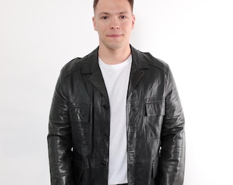 May include: Black leather jacket with a button-up front, two chest pockets, and two lower pockets. The jacket is worn over a white t-shirt and black pants. The jacket has a classic, vintage style.