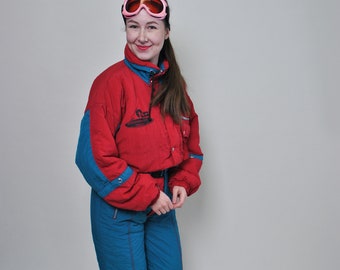 May include: A vintage ski suit in red and teal. The jacket has teal accents on the sleeves and collar, and the pants are teal. The suit is worn with pink ski goggles. A retro winter sports outfit.