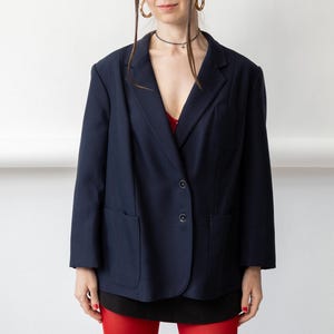 Puede incluir: Una americana azul marino con solapa de muesca, dos botones frontales, un bolsillo en el pecho y dos bolsillos de parche inferiores. La prenda se lleva sobre un top rojo y una falda negra, combinada con medias de color rojo brillante y aros dorados.