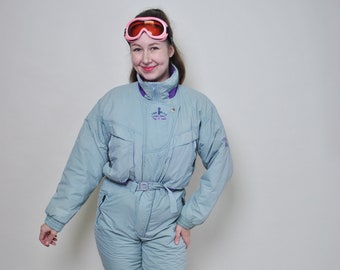 May include: A light blue ski suit with a purple zipper and a white logo that says "The Village" on the chest. The suit has a belt and is worn with white sneakers and pink ski goggles.