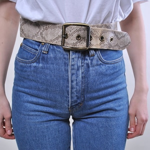 Może przedstawiać: Jasnobrązowy pasek ze skóry węża z pozłacaną klamrą i metalowymi oczkami. Pasek jest noszony na białej koszulce i niebieskich dżinsach.