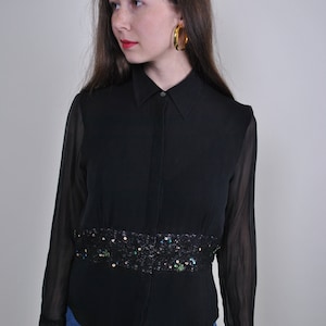 Puede incluir: Camisa negra abotonada con mangas transparentes y una banda decorativa de lentejuelas en la cintura. Los puños están adornados con encaje. La modelo lleva pendientes de aro dorados y vaqueros azules.
