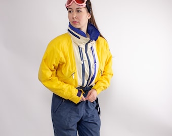 May include: A vintage yellow and navy blue ski suit with a cream-colored upper section and blue accents. The suit features a high collar, zippered pockets, and a belt. The person is wearing pink ski goggles.