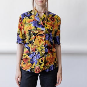 May include: A short-sleeved, double-breasted blouse featuring a vibrant floral pattern in shades of yellow, purple, orange, and black. The garment includes a notched collar, two front flap pockets, and six round, gold-toned buttons down the front.