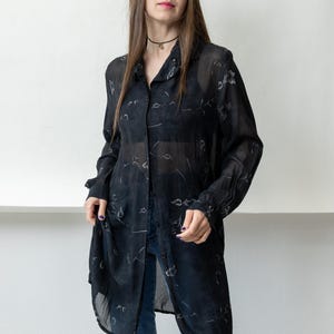 May include: A black, sheer button-down shirt with a floral pattern. The long-sleeved shirt is worn over a dark top and paired with blue jeans. The shirt is open, revealing the outfit underneath. The model is standing in front of a white wall.