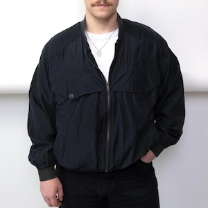 Könnte beinhalten: Dunkelblaue Bomberjacke mit Reißverschluss, zwei Vordertaschen und gerippten Bündchen. Die Jacke wird über einem weißen T-Shirt und einer schwarzen Hose getragen. Die Person steht vor einem weißen Hintergrund.