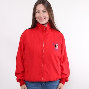 Puede incluir: Chaqueta roja brillante con cremallera, cuello alto y puños elásticos. La chaqueta presenta un pequeño emblema bordado en el pecho izquierdo. El modelo lleva vaqueros azules.