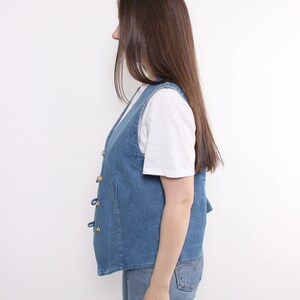 Op de afbeelding: Een blauwe denim vest met gouden knopen en een wit T-shirt eronder.