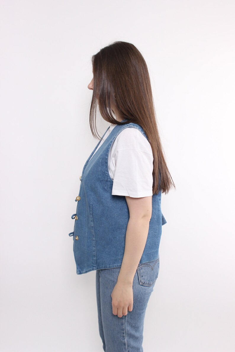 Op de afbeelding: Een blauwe denim vest met gouden knopen en een wit T-shirt eronder.