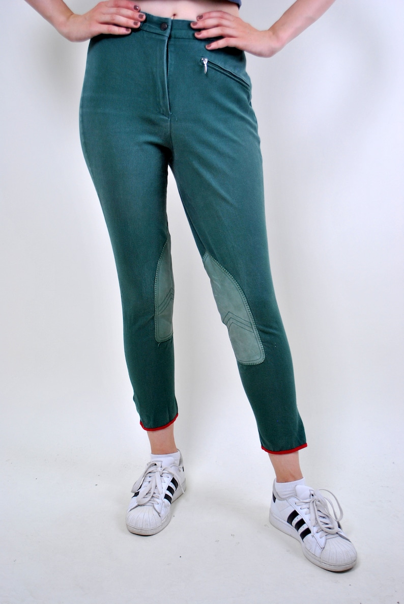 May include: Green riding pants with knee patches and red trim at the bottom. The pants are worn with white sneakers with black stripes.
