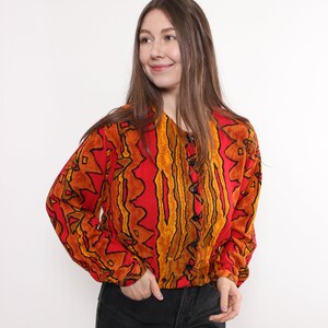May include: A long-sleeved, button-down blouse with a vibrant red background and an abstract pattern in shades of orange and black. The blouse is paired with black denim jeans. The model is smiling.