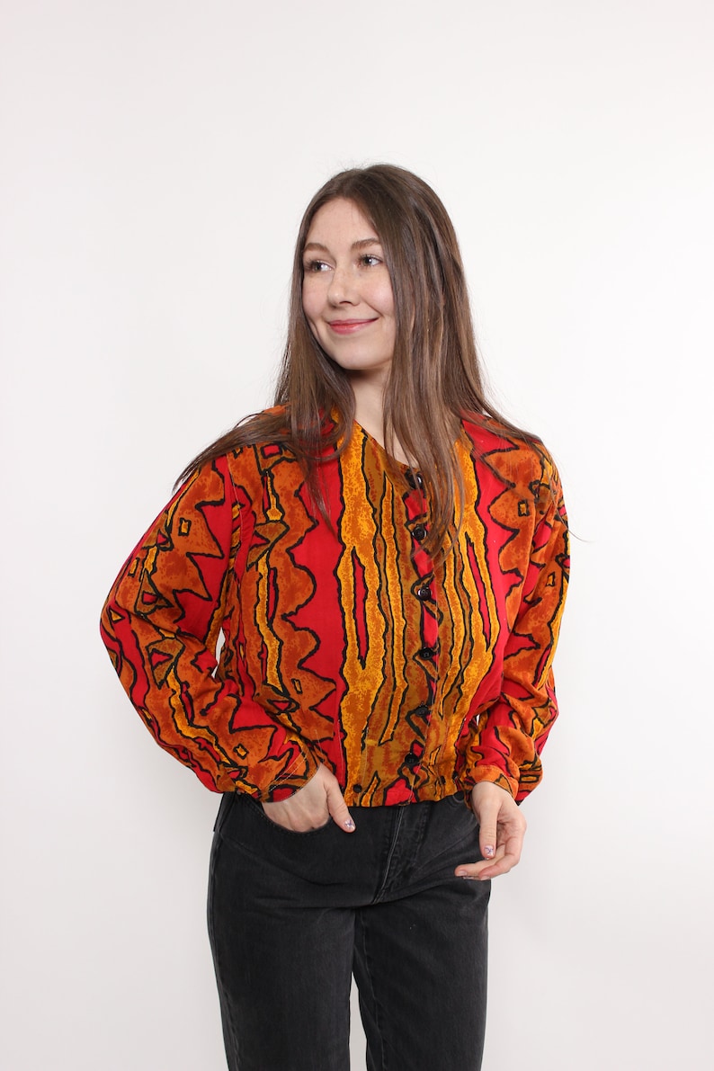 May include: A long-sleeved, button-down blouse with a vibrant red background and an abstract pattern in shades of orange and black. The blouse is paired with black denim jeans. The model is smiling.