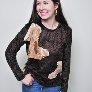 May include: A woman wearing a brown velvet long-sleeved shirt with beige fabric appliques. The shirt has a round neckline. She is also wearing blue jeans.