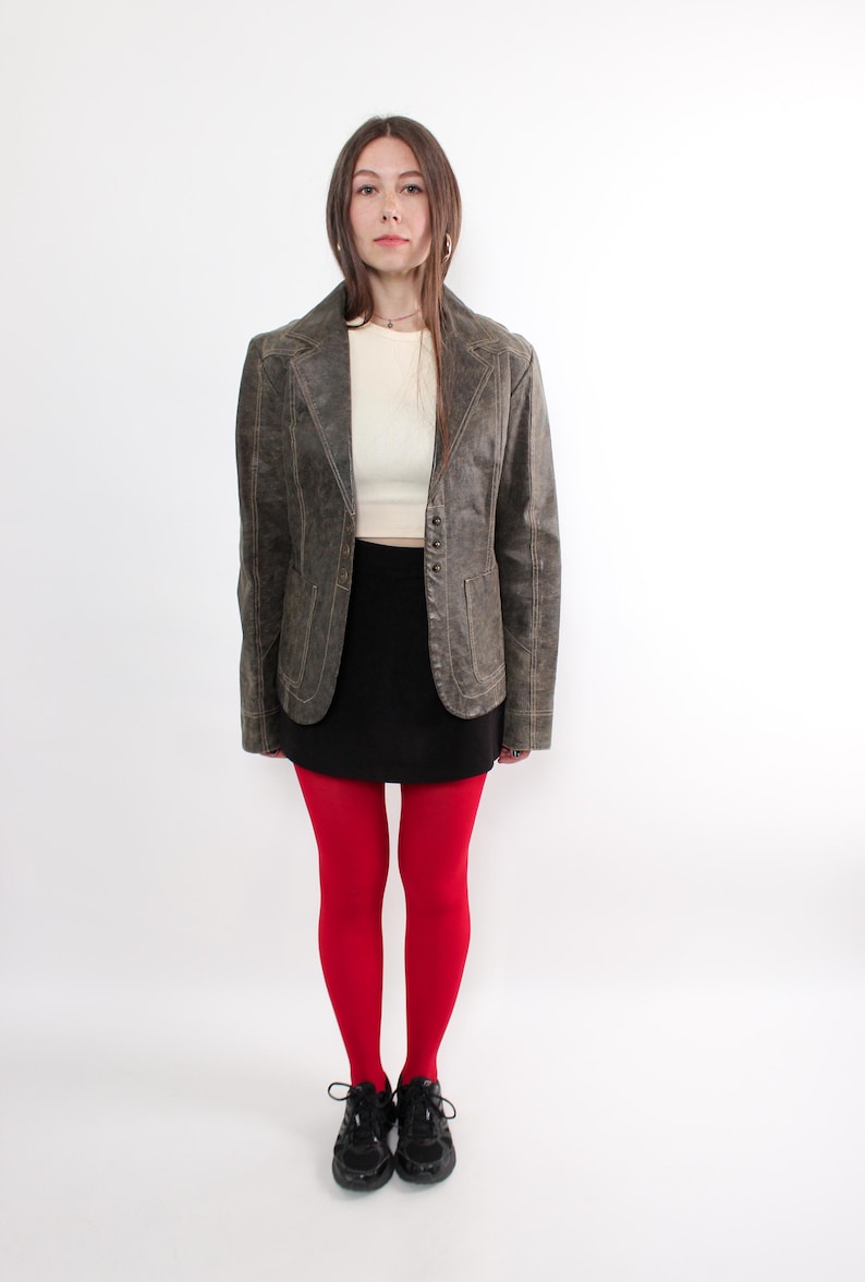Puede incluir: Chaqueta de cuero marr&oacute;n de estilo vintage con solapas y cierre de botones. El conjunto incluye una camiseta crema, una falda negra, medias rojas brillantes y zapatillas negras. El conjunto se presenta sobre un fondo blanco.