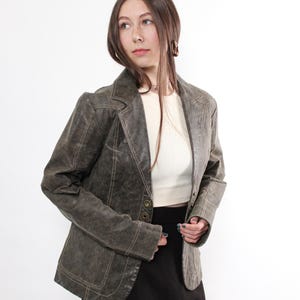 Puede incluir: Chaqueta blazer verde oliva de estilo vintage con solapa de muesca y cierre de bot&oacute;n. La chaqueta presenta costuras visibles y se combina con una camiseta blanca, una falda negra y medias rojas, sobre fondo blanco.