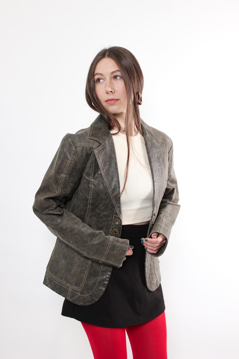 Puede incluir: Chaqueta blazer verde oliva de estilo vintage con solapa de muesca y cierre de bot&oacute;n. La chaqueta presenta costuras visibles y se combina con una camiseta blanca, una falda negra y medias rojas, sobre fondo blanco.