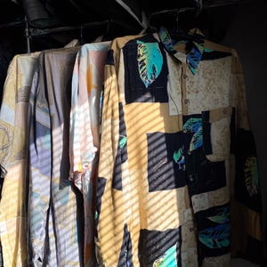 May include: A collection of vintage button-down shirts hanging on a rack. The shirts feature various patterns and colors, including beige, blue, and yellow. One shirt has a leaf design with black and teal accents. The sunlight casts shadows across the garments.