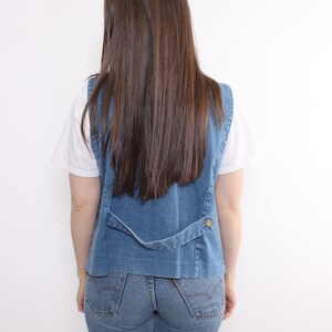 Op de afbeelding: Een blauwe denim vest met een knoopsluiting en een wit T-shirt eronder. De persoon draagt ​​een blauwe jeans.