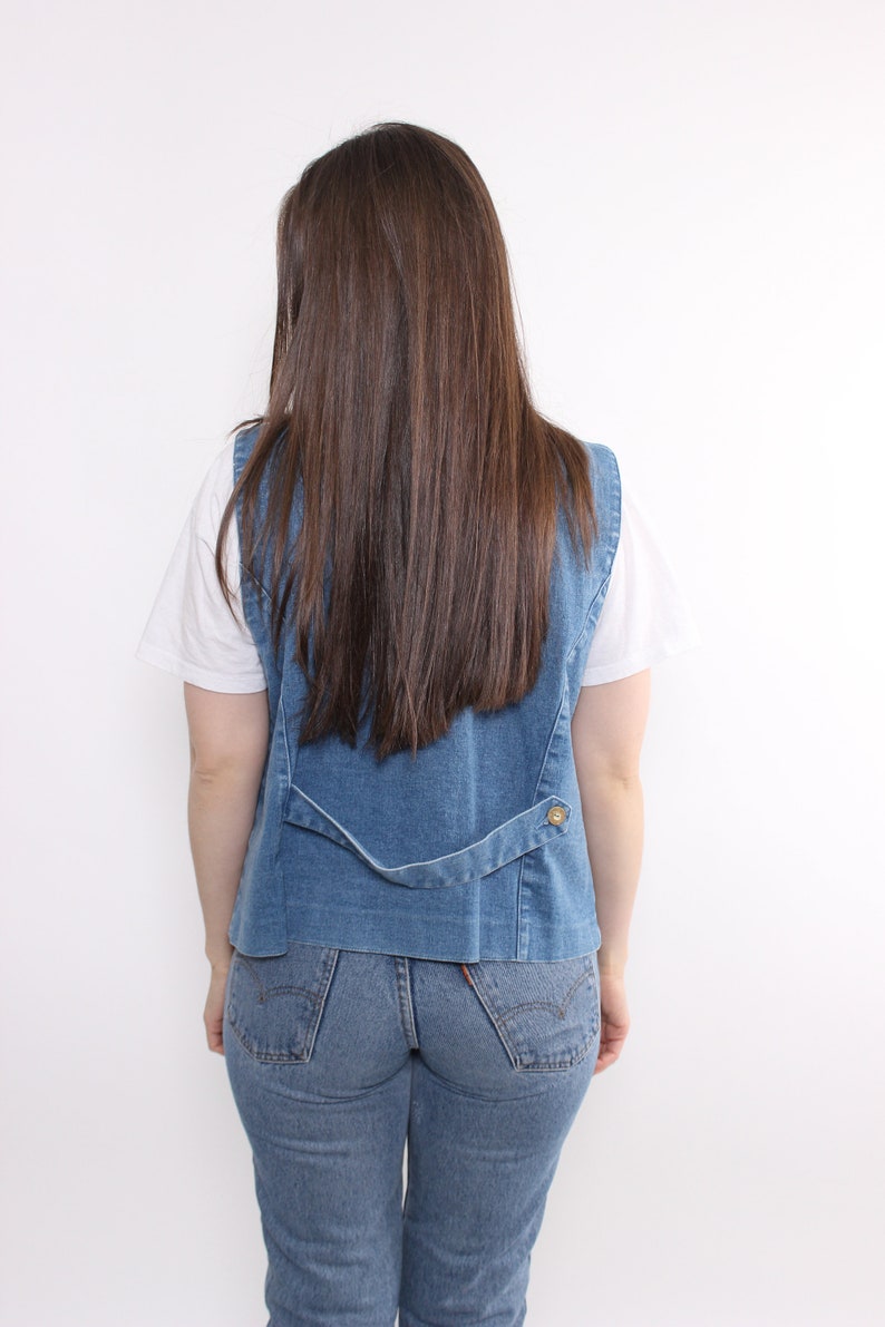 Op de afbeelding: Een blauwe denim vest met een knoopsluiting en een wit T-shirt eronder. De persoon draagt ​​een blauwe jeans.