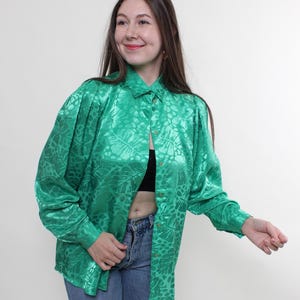 May include: A bright green button-down shirt with a leaf pattern. The long-sleeved shirt has a collared neckline and gold-colored buttons. The fabric has a shiny, satin-like appearance. The shirt is paired with blue jeans.