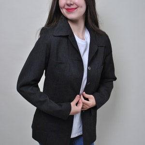 Pode incluir: Uma mulher vestindo um blazer preto com uma camisa branca por baixo. Ela está usando jeans azul e tem as mãos juntas na frente dela.