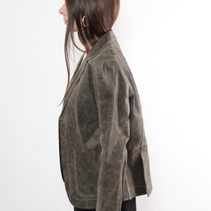 Puede incluir: Blazer verde oliva de estilo vintage con acabado texturizado. La chaqueta presenta solapas con muescas, cierres de botones y bolsillos delanteros. El blazer se usa con una falda negra y medias rojas, creando un look en capas.