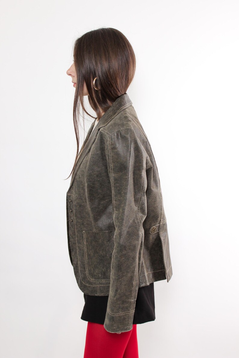Puede incluir: Blazer verde oliva de estilo vintage con acabado texturizado. La chaqueta presenta solapas con muescas, cierres de botones y bolsillos delanteros. El blazer se usa con una falda negra y medias rojas, creando un look en capas.