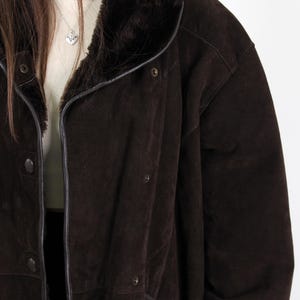 Puede incluir: Un abrigo de ante marr&oacute;n oscuro con cuello y forro de piel sint&eacute;tica. El abrigo tiene cierre de botones, bolsillos y ribetes de cuero. El abrigo es de estilo vintage.