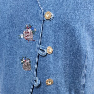 Op de afbeelding: Een blauwe denim vest met knoopsluiting en geborduurde bloemenmotieven. De vest is gemaakt van lichtblauwe denim stof en heeft houten knopen.