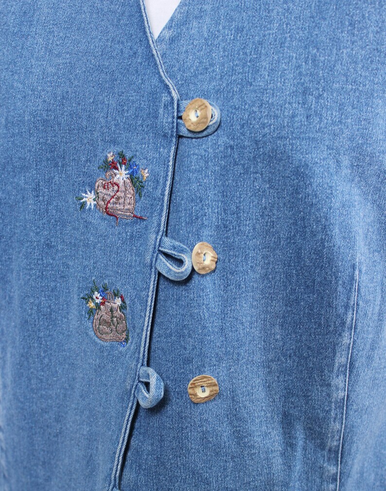 Op de afbeelding: Een blauwe denim vest met knoopsluiting en geborduurde bloemenmotieven. De vest is gemaakt van lichtblauwe denim stof en heeft houten knopen.